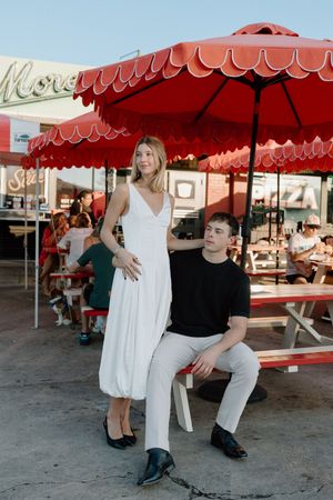 Couple sitting and posing casually under red market umbrellas in an outdoor setting.