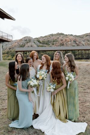 A wedding party in sage green and blue dresses laughs together while holding white floral bouquets outdoors.