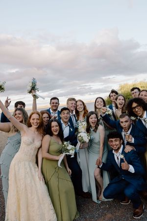 A joyful wedding party in gray suits and olive dresses poses together outdoors at sunset.