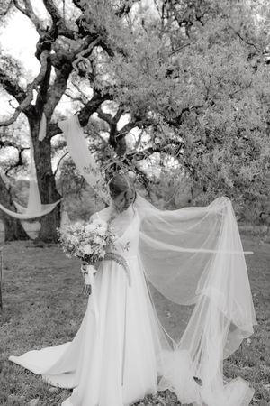 A bride in a flowing white dress and long veil stands beneath oak trees holding a bouquet in black and white photography.