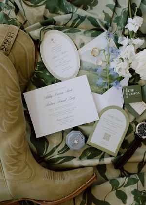 Elegant wedding invitation suite with white cards and envelopes arranged among green foliage and cowboy boots.