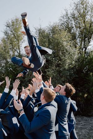 Groomsmen in navy suits toss a groom into the air during a fun wedding photo sequence outdoors.