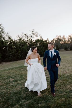 Newlyweds walk together through a grassy field at sunset, with the bride holding her white gown as they stroll.