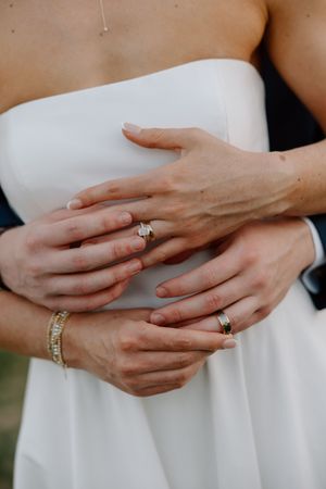Close-up of hands wearing elegant diamond rings and jewelry against a white wedding dress.