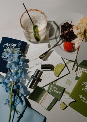 Flatlay of blue delphinium flowers, cocktail glasses, and event stationery with lime and strawberry garnishes.