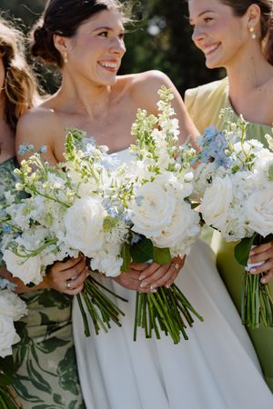Bridesmaids in white dresses hold elegant white floral bouquets while smiling together outdoors in natural sunlight.