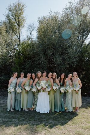 Wedding party in sage green and cream colored dresses poses together against a backdrop of trees and natural scenery.