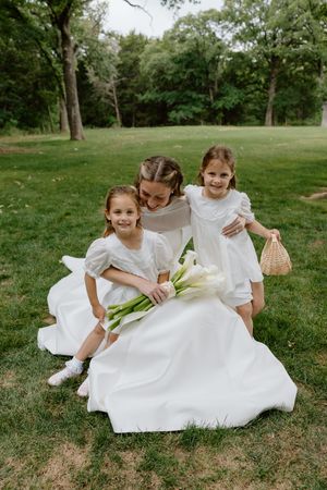 Bride in white gown hugs two flower girls in white dresses holding calla lilies in a lush green park.
