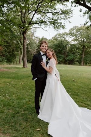 Bride in elegant white gown embraces groom in black tuxedo outdoors in a lush green park setting.