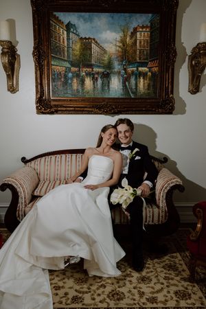 Bride and groom sit together on ornate striped sofa beneath a classic oil painting in elegant venue.