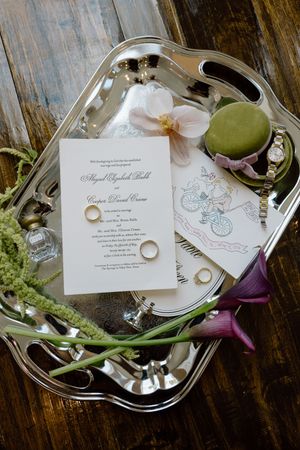 Silver tray with wedding invitation, gold rings, macarons, floral accents, and a watch on rustic wood.