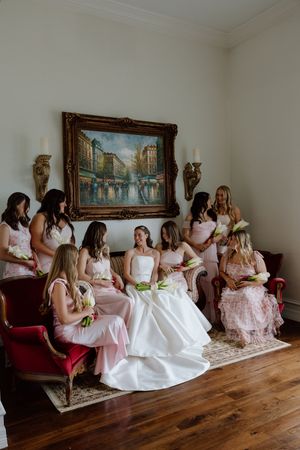 Bride in white gown sits with bridesmaids in pink dresses, holding bouquets in an elegant room with a Paris painting.
