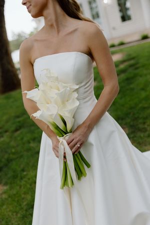Bride in strapless white gown holding elegant calla lily bouquet tied with ribbon, standing outdoors on green lawn.