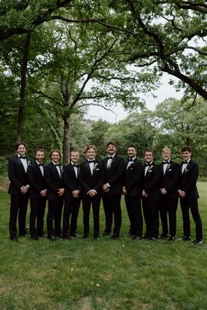 Ten groomsmen in black tuxedos with bow ties pose outdoors on green grass surrounded by lush trees.