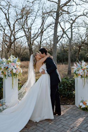 A romantic outdoor wedding ceremony with white draped columns and colorful floral arrangements lining a stone pathway.