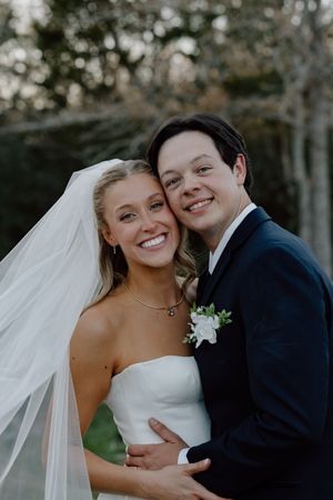 A happy couple embraces on their wedding day, with the bride wearing a white gown and veil against a natural outdoor backdrop.