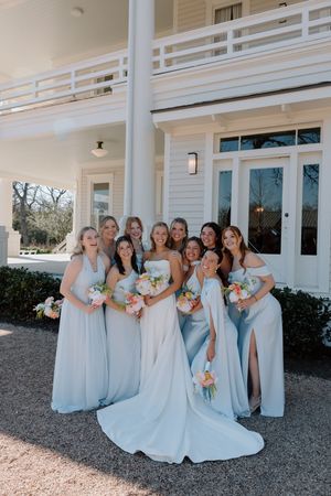 Wedding party wearing light blue bridesmaid dresses pose together outside a white building.