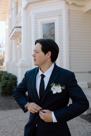 A groom in a classic navy suit with white boutonniere adjusts his jacket while standing outside a white Victorian building.