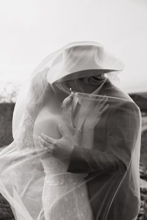 A romantic black and white wedding photo showing a couple embracing under a flowing veil.
