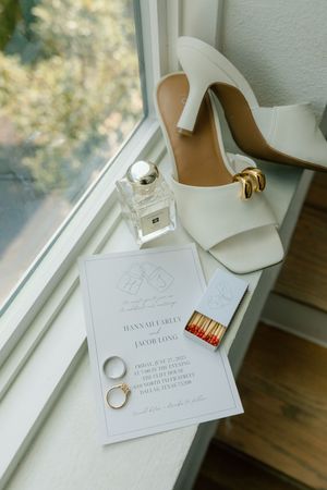 Wedding invitation suite with white heels, matches, and a ring displayed elegantly by a window.