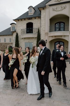 A wedding party in black attire and white gown gather outside an elegant stone mansion with French architectural details.