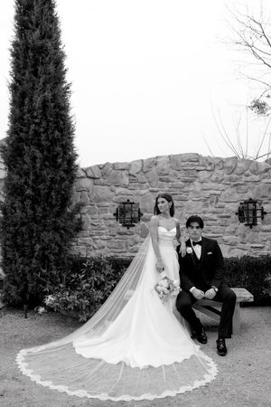 A wedding couple sits together on a stone wall in a garden with evergreen trees and a flowing white dress.