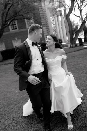 Black and white photo of a smiling bride and groom walking together on grass outside a brick building with columns.