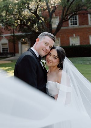 A bride and groom pose romantically outdoors, her flowing veil billowing, with a brick building backdrop.