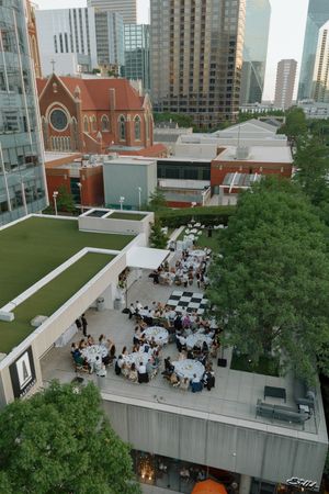 Aerial view of an outdoor rooftop event with round tables and guests, surrounded by city buildings and a historic red brick church.