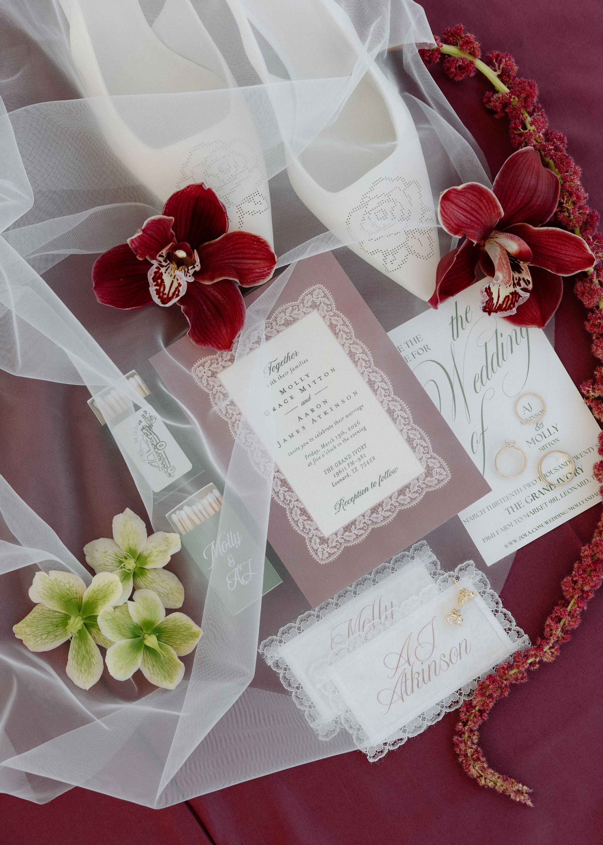 Elegant wedding flat lay with burgundy orchids, lace invitations, white heels, and floral details on a deep red backdrop.