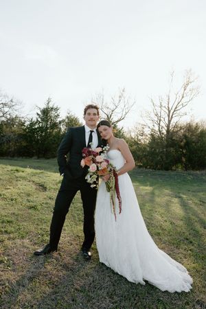 A bride and groom pose together outdoors in a grassy field, she holds a colorful bouquet in her white gown.