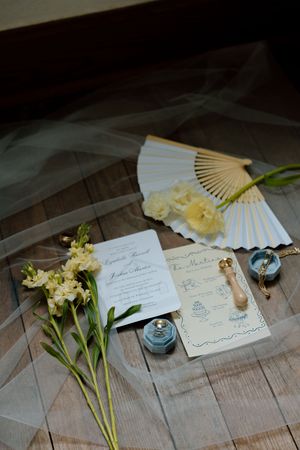 Wedding invitation details with yellow flowers, decorative fan, and blue accessories arranged on wooden surface.