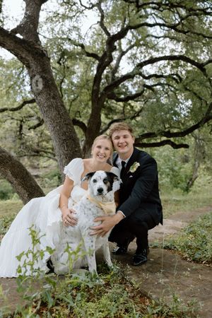 A couple in wedding attire sits on the ground outdoors with their white dog under large oak trees.