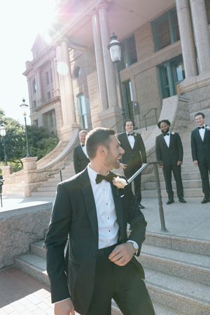 Groomsmen in black tuxedos and white shirts pose playfully on outdoor stone steps of a grand building.