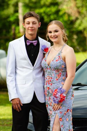Couple in formal attire posing for prom photos outdoors with white tuxedo and floral dress.