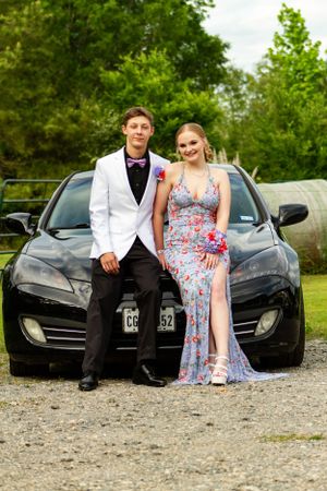 Formal couple posing against a black car on a gravel path.