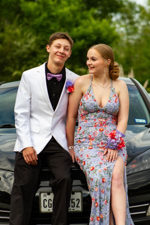Couple in formal wear smiling while sitting on car hood outdoors.