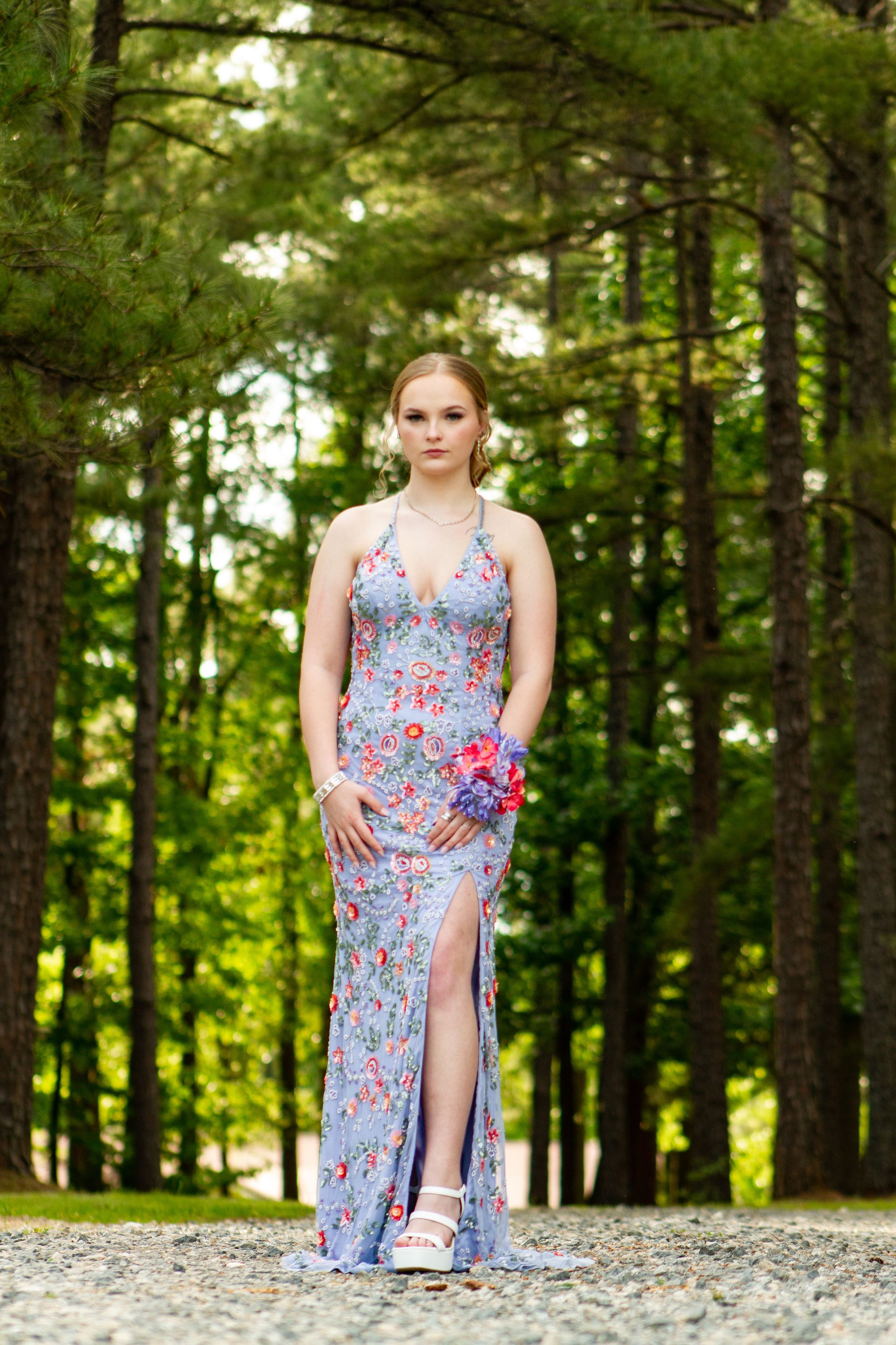 Woman in blue floral gown posing on gravel path in pine forest.
