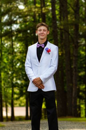 Young man in white tuxedo jacket and bow tie posing in forest setting.