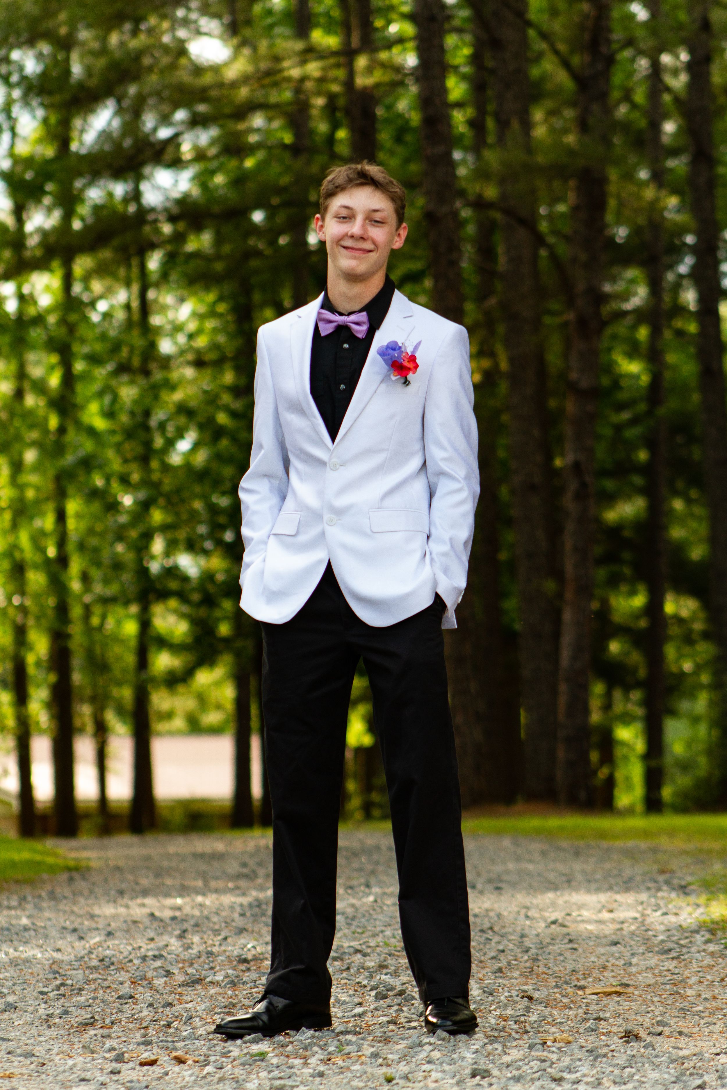Young man in white dinner jacket smiling in wooded outdoor setting.