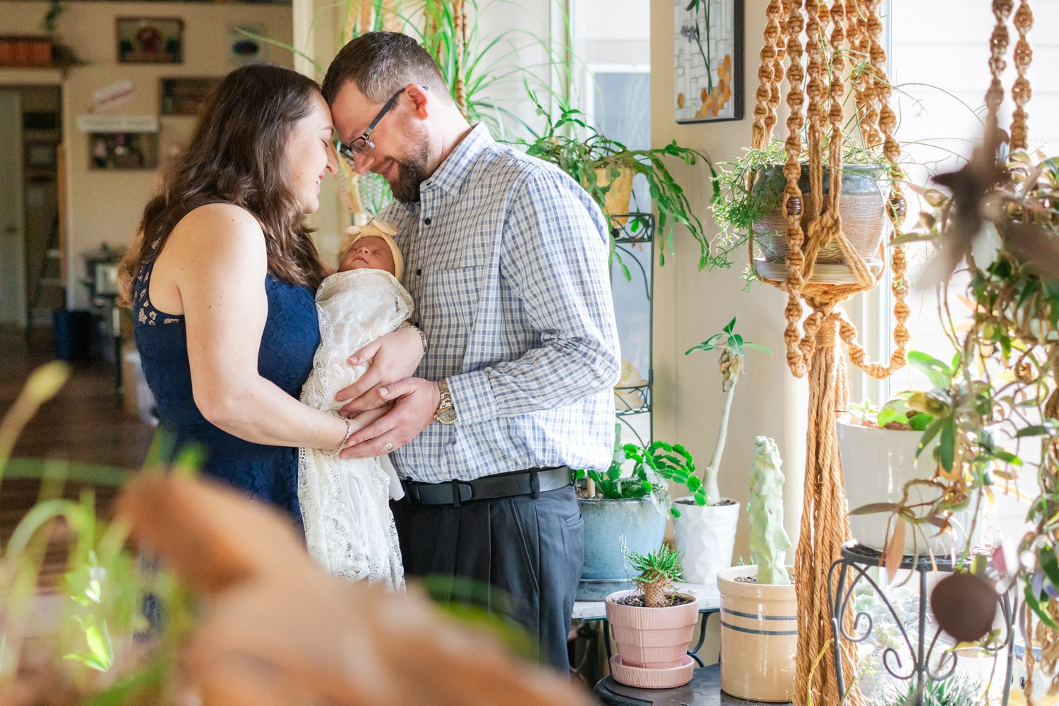 Mom and dad embrace holding newborn daughter by the window in a sunroom with lots of plants