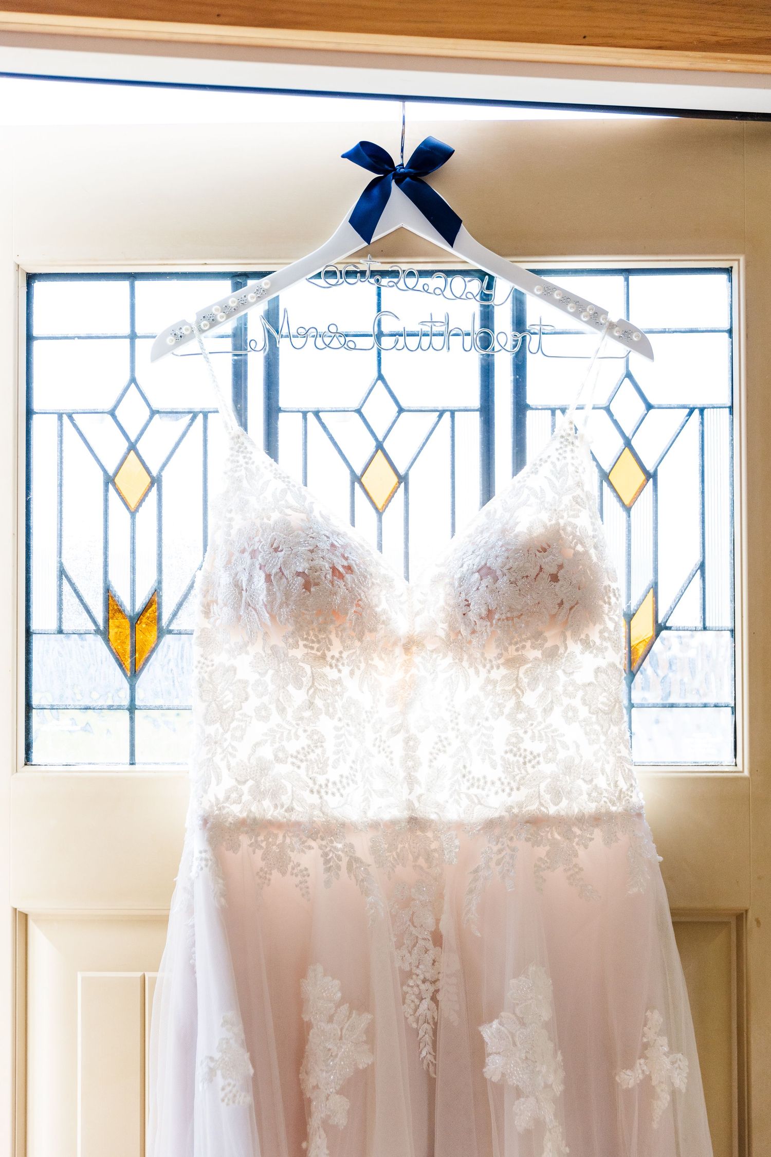 White wedding dress hangs elegantly against stained glass window with blue bow accent.