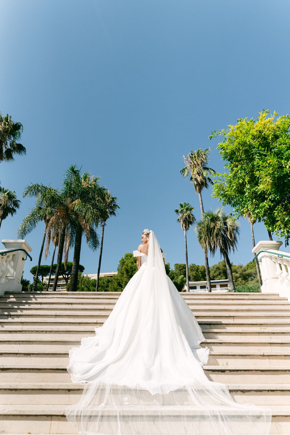 A flowing white wedding dress with long train displayed on grand outdoor stone steps surrounded by palm trees against blue sky.
