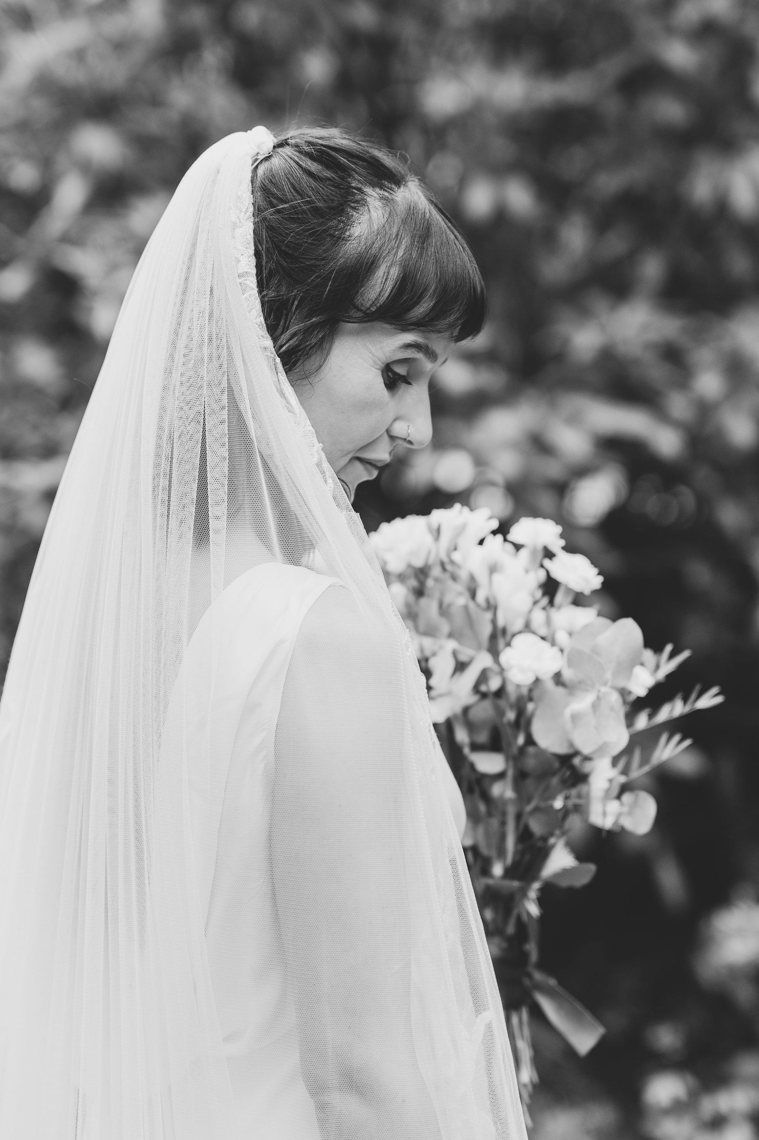 A bride in a flowing white veil and dress holds a bouquet of white flowers in a black and white photograph.