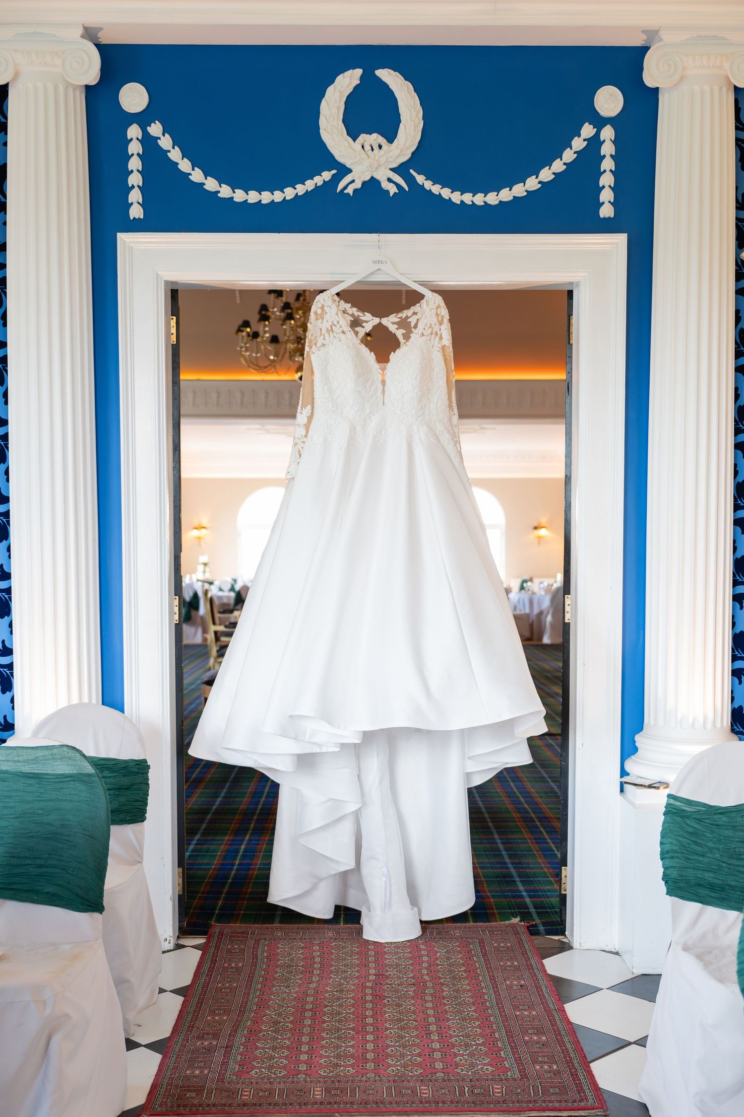 Elegant white lace wedding dress with long sleeves hanging in a blue and white decorative archway entrance.