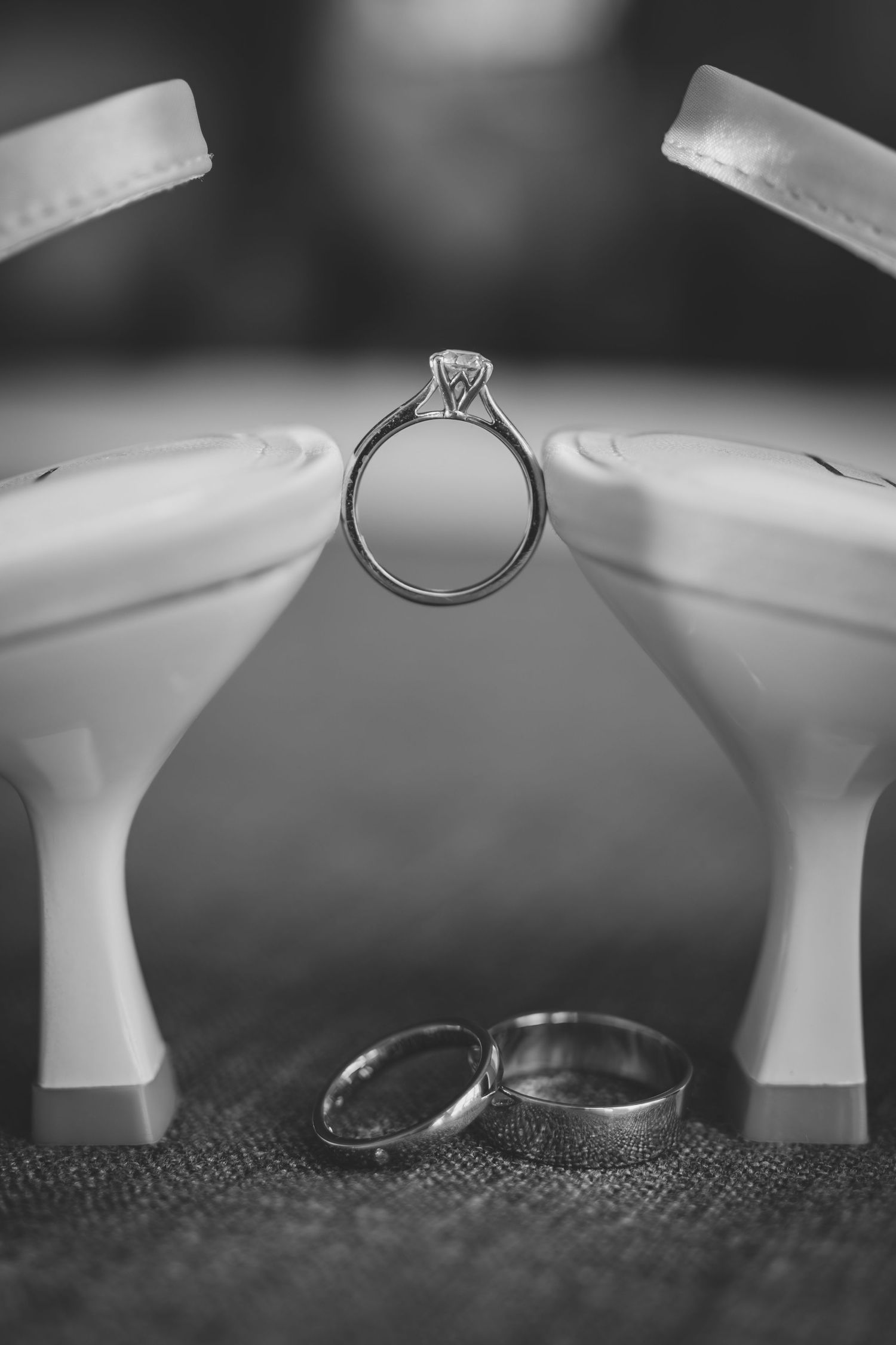 Black and white photo of engagement and wedding rings artistically arranged between two white high heel shoes.