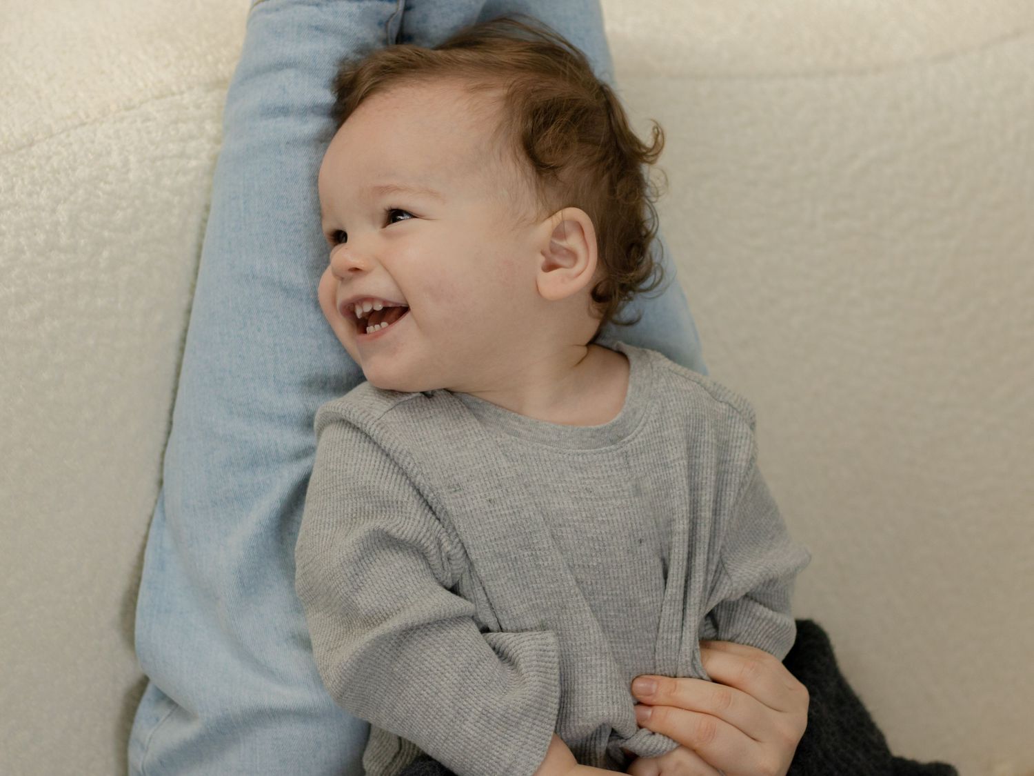 Toddler in a gray sweater laughing while lying on a light blue surface.