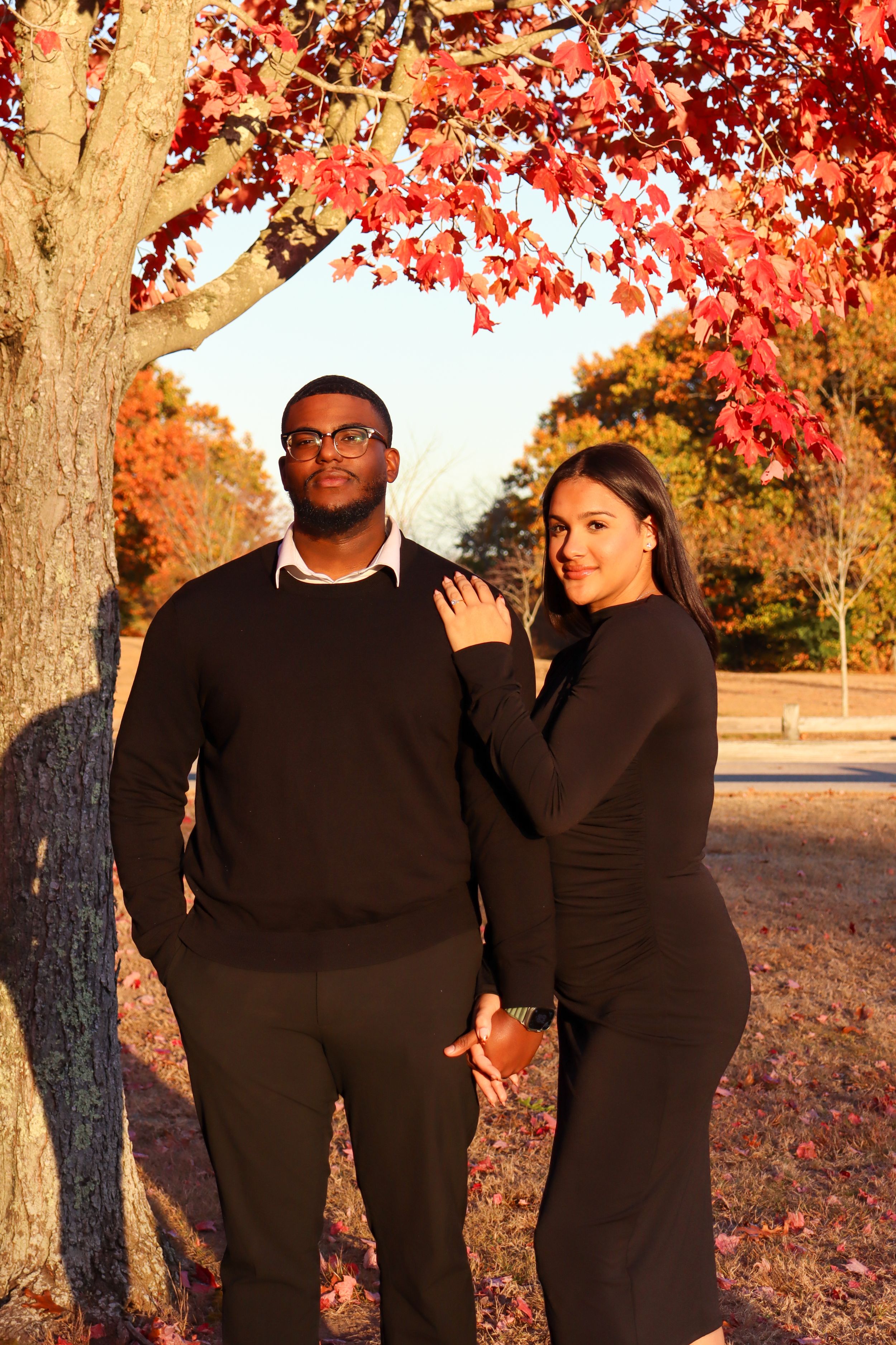 A couple poses together under red fall foliage in matching black outfits.