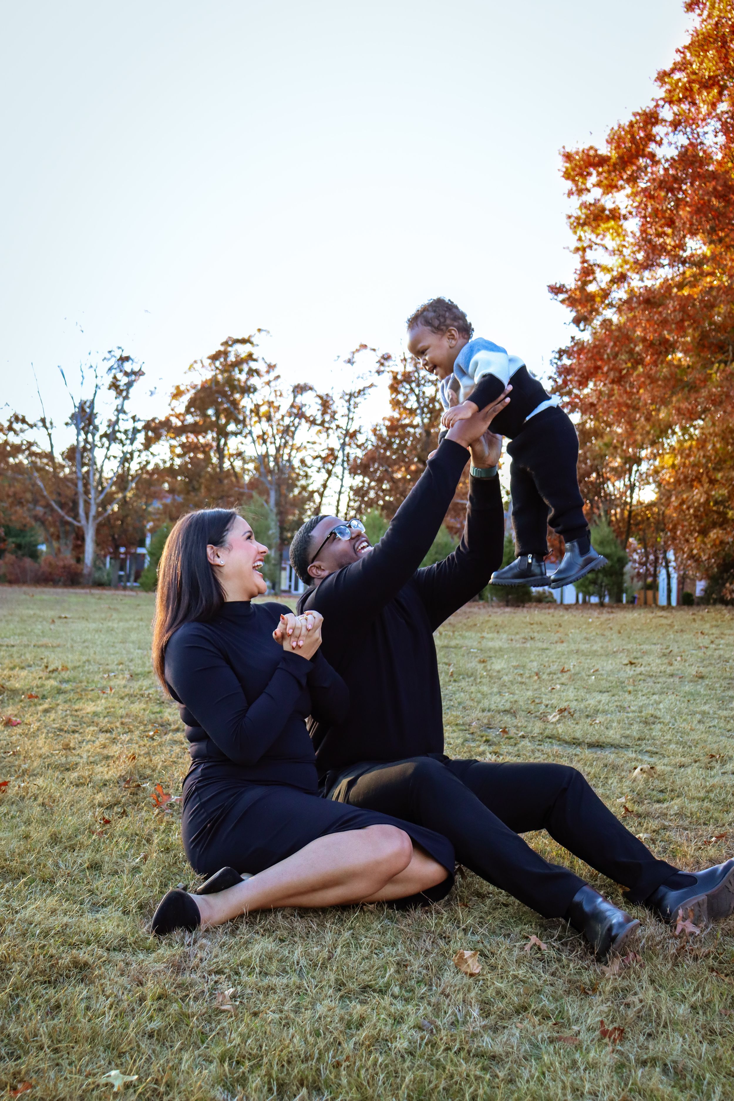 A playful family moment captured on grass with fall trees in the background.