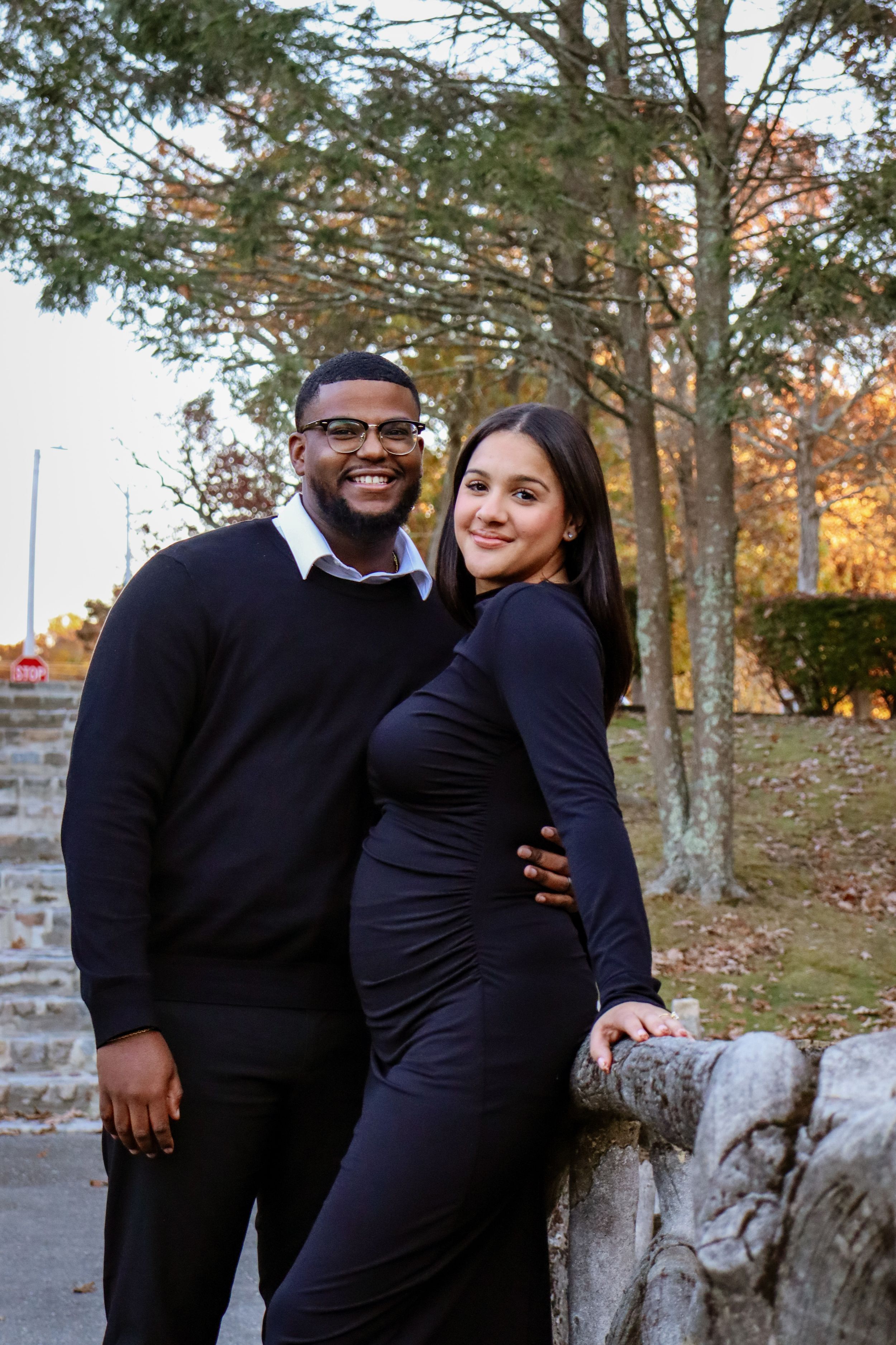 A couple poses against a stone wall with tall pine trees in the background.
